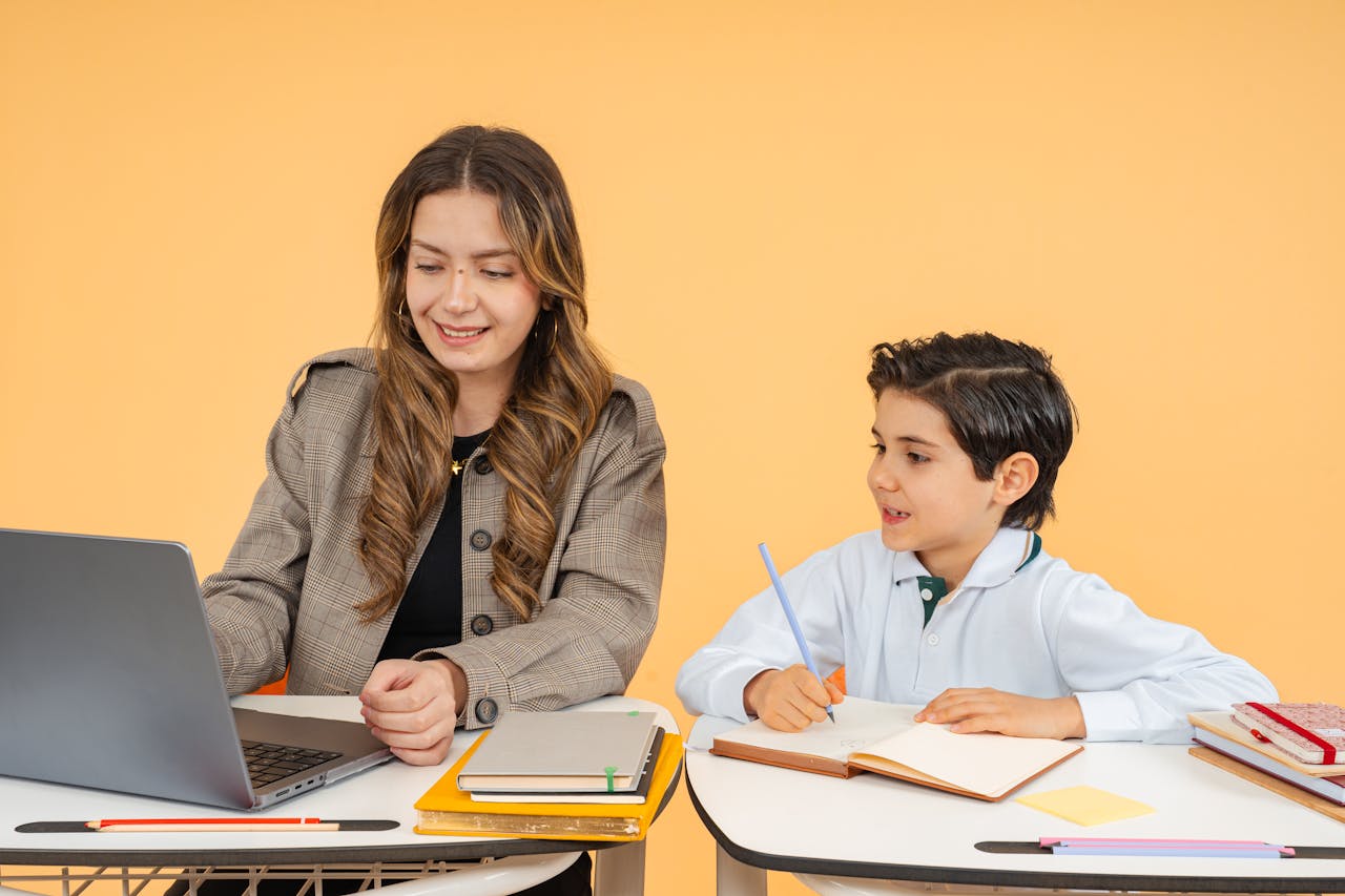 A female teacher helps a young student with his studies using a laptop in a classroom setting.